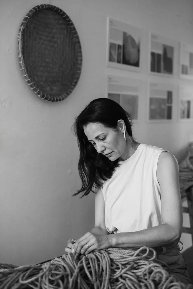Black and white photograph of Angelita Alves working, with her grandfather's woven sieve hanging on the wall behind her